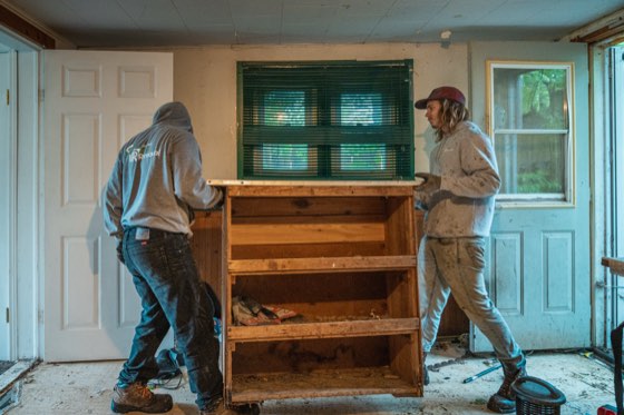 Two crew members carrying a large wooden hutch out through the front door of a house