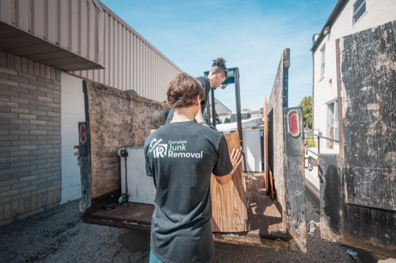 A crew member in a Complete Junk Removal shirt loading salvaged boards into the trailer beside a building